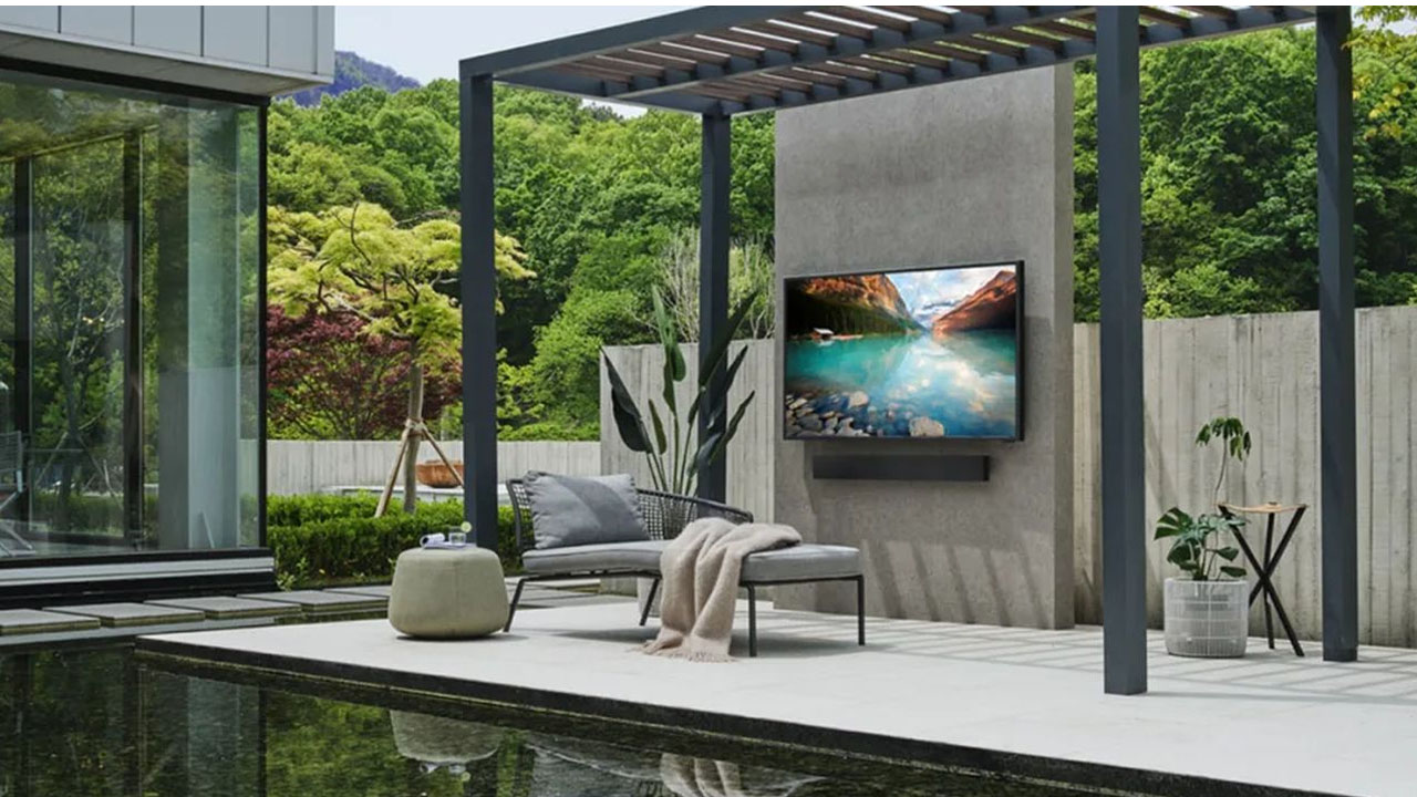 An outdoor terrace setup featuring a large Samsung outdoor TV mounted on a wall, surrounded by comfortable seating and subtle ambient lighting for alfresco entertainment.