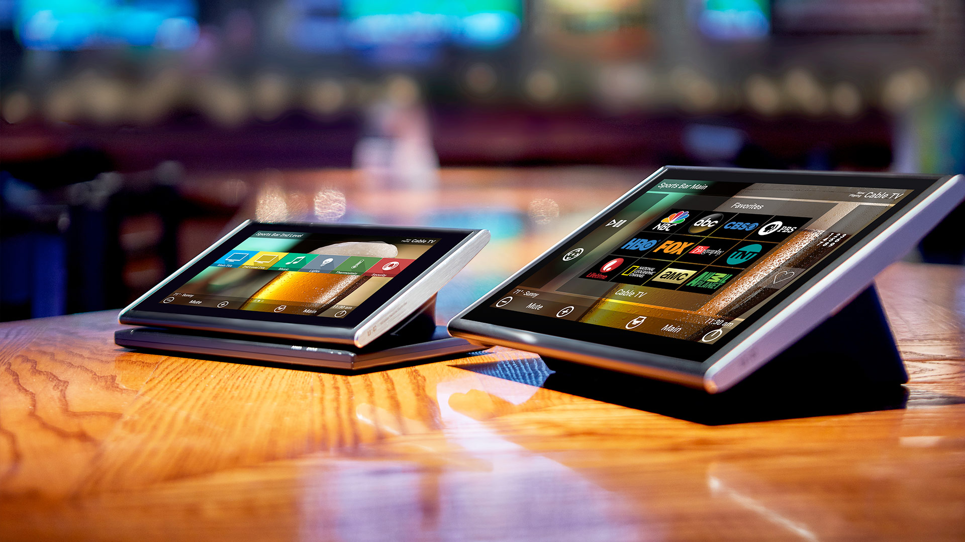Two tabletop touchscreens rest on a polished bar, each showing colorful control interfaces for TV channels, lighting, and audio in a lively sports-bar setting.