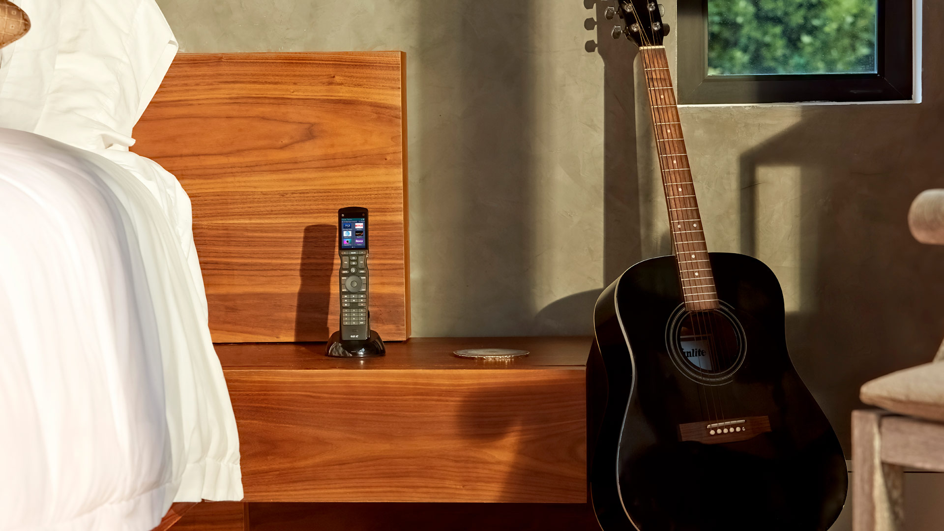 Cozy bedroom detail: black acoustic guitar leans against a concrete wall next to a wooden nightstand where an upright touchscreen universal remote rests in its charging dock; white bedding is visible on the left edge of the frame.