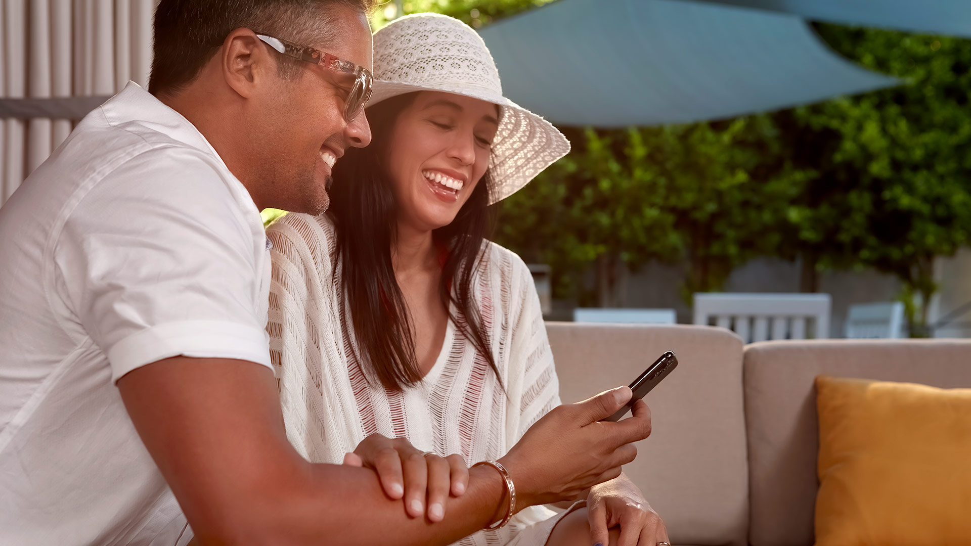 Close-up of the same couple smiling and laughing while looking at a smartphone on a shaded patio sofa, with blurred green foliage and outdoor furniture in the background.