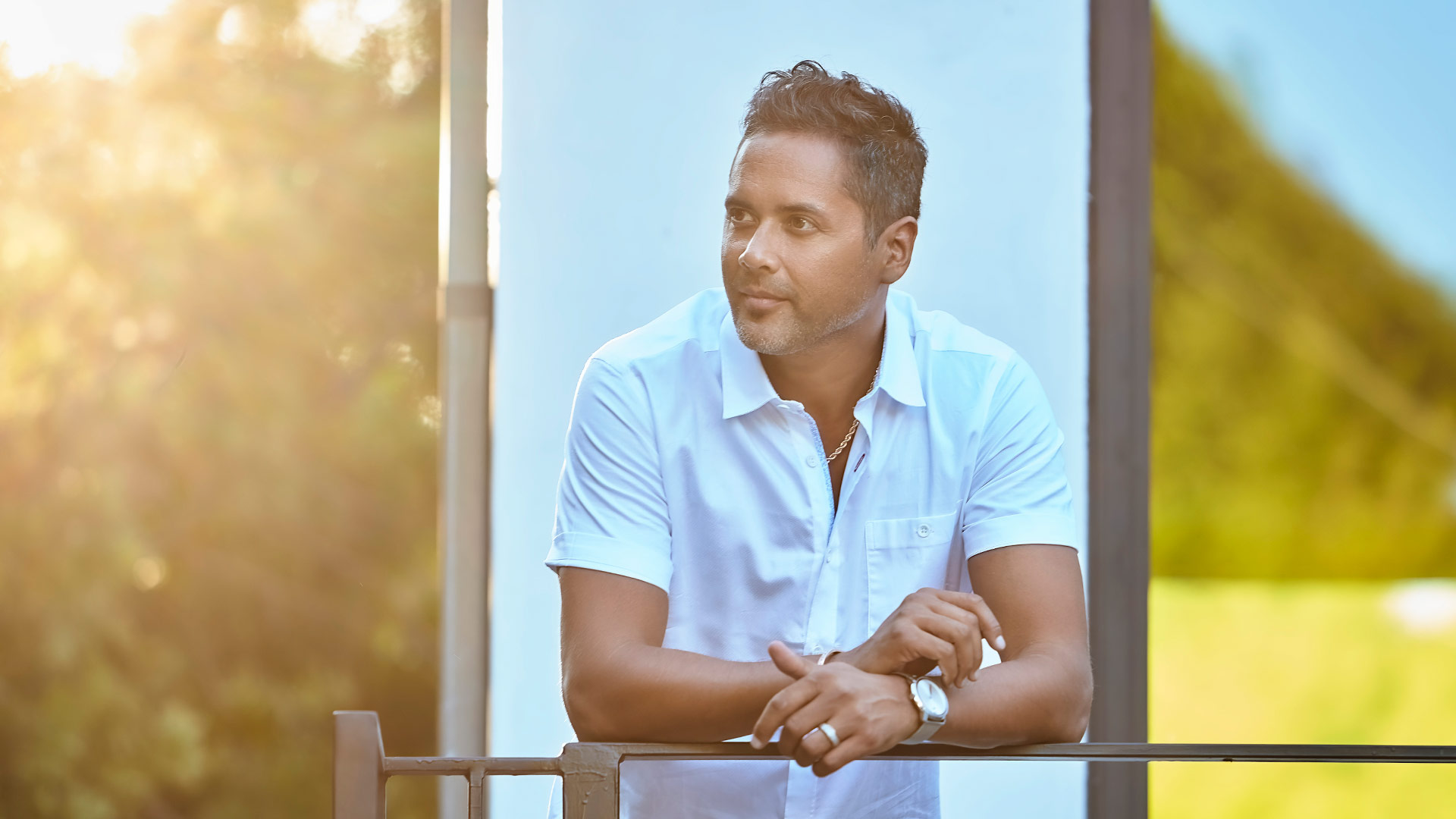 Sun-lit portrait of the man leaning casually on a metal deck railing, gazing thoughtfully into the distance while wearing a white collared shirt, watch, and gold chain.