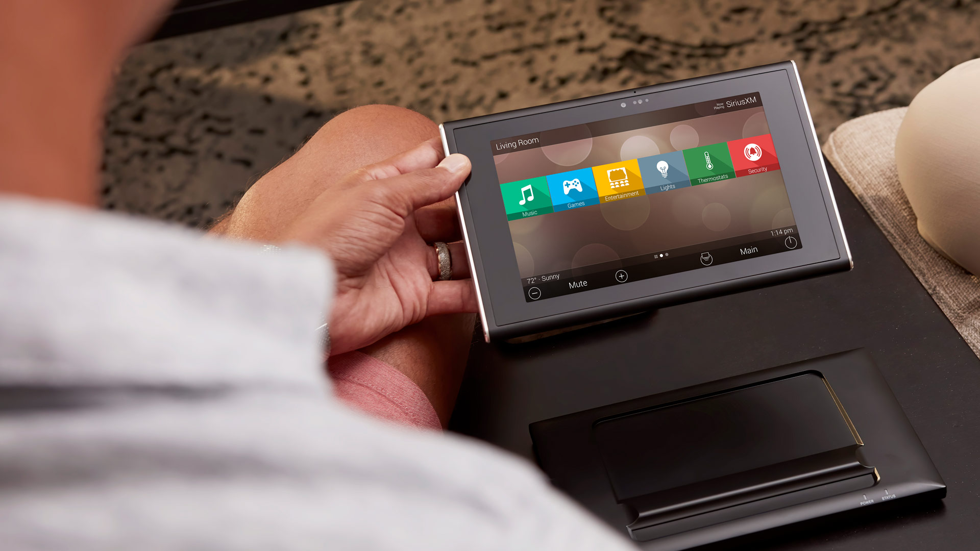 Close-up over someone’s shoulder of a handheld smart-home tablet displaying icons for music, games, lights, and security; the charging dock sits on a nearby side table.