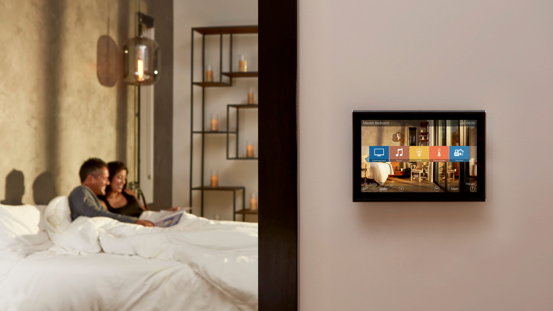 Wall-mounted touch panel showing master-bedroom controls, while a smiling couple relaxes in bed in the softly lit background of the room.