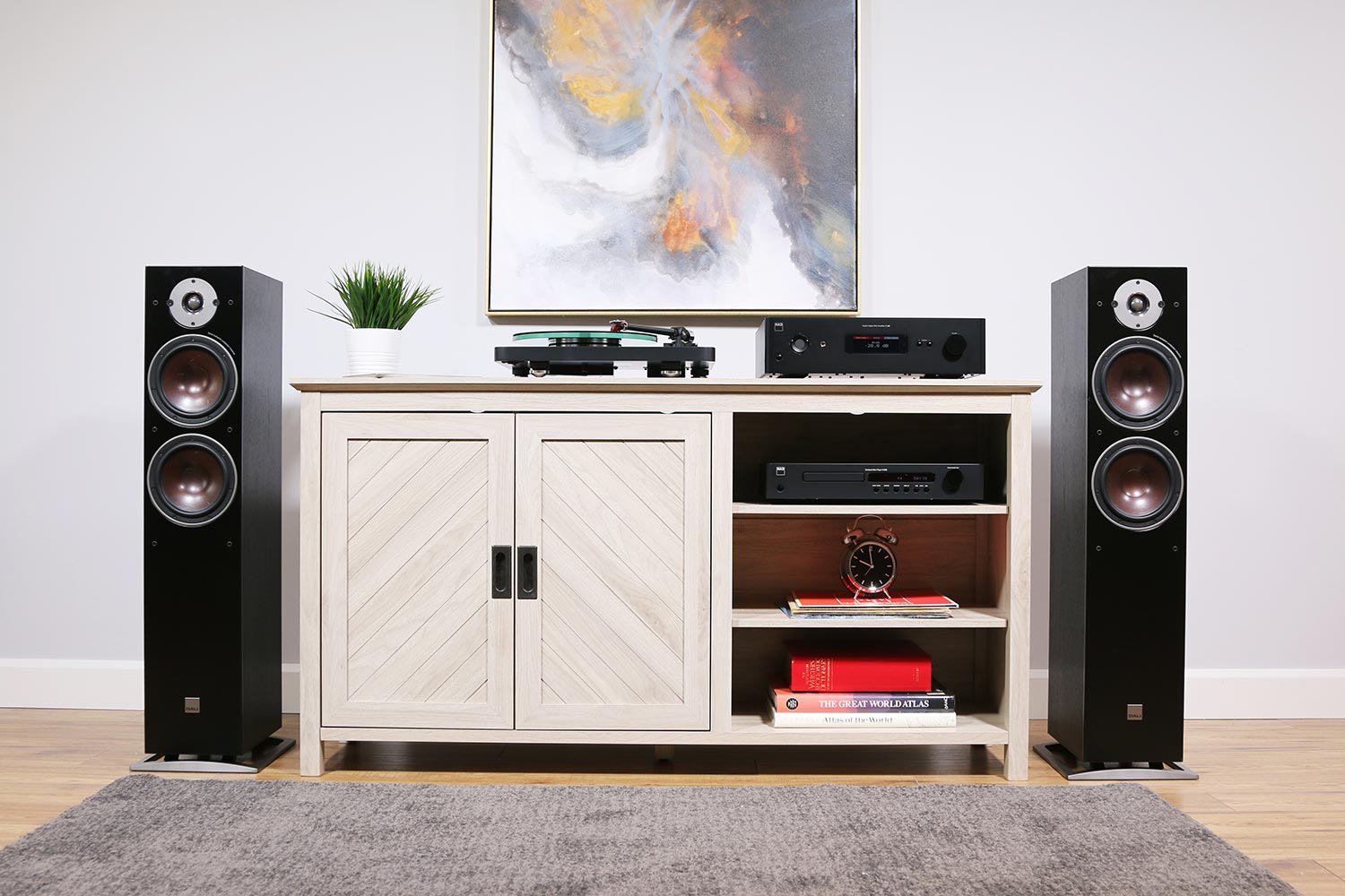 Light wood media console with chevron doors is flanked by two black floor-standing speakers; a glass-platter turntable and a black NAD C 388 integrated amplifier sit on top, creating a modern stereo setup beneath a colorful abstract painting.